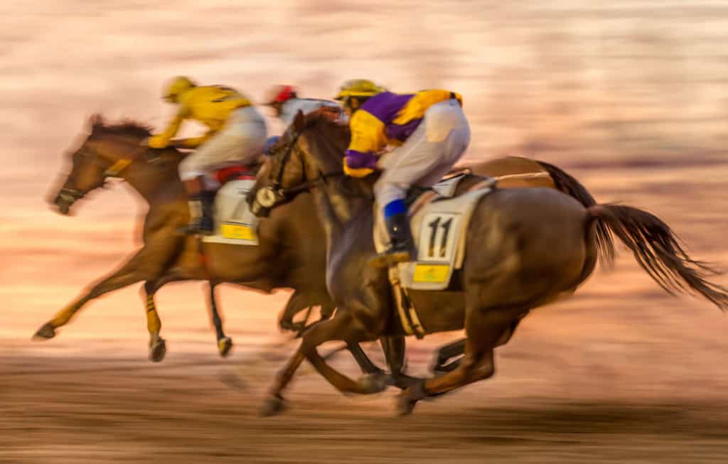 Três cavalos de corrida avançando rapidamente com jóqueis montados, mostrando movimento intenso e velocidade em uma pista sob luz dourada