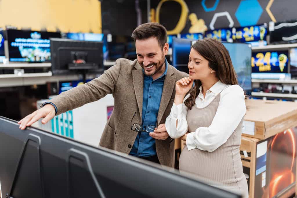 Um homem e uma mlher observando uma televisão; o homem sorri e a mulher está com uma das mãos no queixo